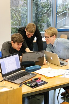 Foto von drei Studenten. Sie sitzen um einen Tisch herum und arbeiten an ihren Laptops. Auf dem Tisch liegen weitere Arbeitsmaterialien wie Stifte und Blätter.