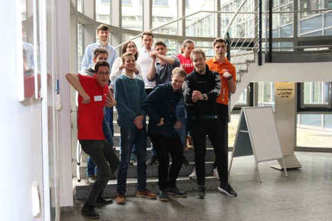 Foto von einer Gruppe Studierender, die vor und teilweise auf einer Treppe stehen und in die Kamera blicken.