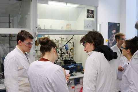Foto von vier Studierenden, die mit Laborkitteln in einem Labor stehen und eine chemische Anlage betrachten.