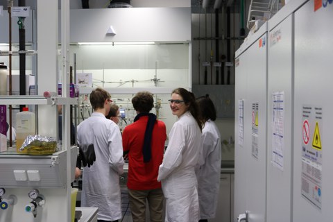 Foto von fünf Studierenden, die mit Laborkitteln in einem Labor stehen und eine chemische Anlage betrachten.
