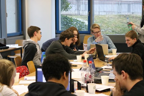Foto von Studierenden während der Teamarbeit. Sie sitzen an einem Tisch und arbeiten an Laptops oder unterhalten sich.