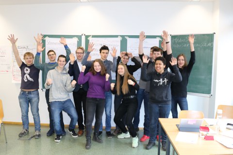 Foto von einer Gruppe Schüler und Schülerinnen während der Projektwoche. Sie stehen vor einer beschriebenen Tafel und schauen direkt in die Kamera. Dabei reisen sie die Hände in die Luft.