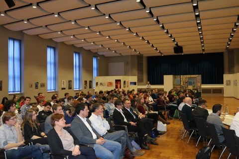 Foto der Abschlussveranstaltung. In einem Saal stehen voll besetzte Stuhlreihen, die seitlich zu sehen sind. Die Personen auf den Stühlen schauen nach vorne in Richtung des Podestes.