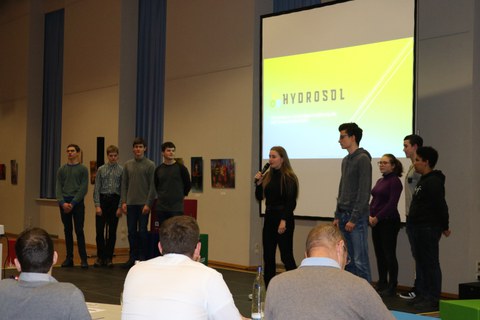Foto einer Abschlusspräsentation einer Gruppe Schüler und Schülerinnen. Sie stehen vor einer Leinwand und eine Schülerin spricht in ein Mikrofon.