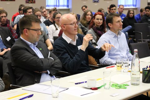 Foto der Jury bei der Abschlussveranstaltung. Drei Männer sitzen an einem Tisch. Der Mann in der Mitte spricht in ein Mikrofon.