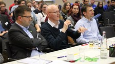 Foto der Jury bei der Abschlussveranstaltung. Drei Männer sitzen an einem Tisch. Der Mann in der Mitte spricht in ein Mikrofon.