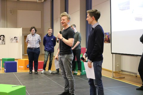 Foto einer Abschlusspräsentation einer Studierendengruppe. Zwei Studenten stehen vor einer Leinwand und einer von beiden spricht in ein Mikrofon. Im Hintergrund ist der Rest der Gruppe zu erkennen, die neben der Leinwand stehen.