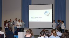Foto einer Abschlusspräsentation einer Studierendengruppe. Die Gruppe steht aufgeteilt jeweils links und rechts neben einer Leinwand.