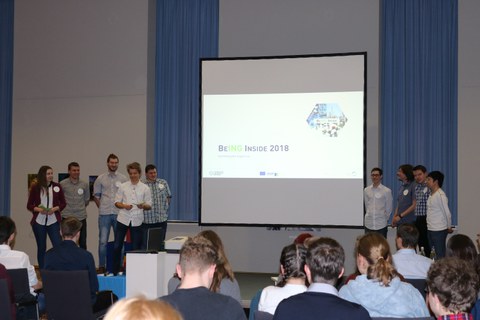 Foto einer Abschlusspräsentation einer Studierendengruppe. Die Gruppe steht aufgeteilt jeweils links und rechts neben einer Leinwand.