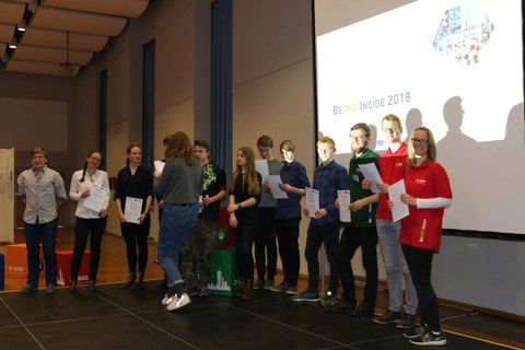 Foto von der Ausgabe der Teilnahmezertifikate. Studierende stehen im Halbkreis vor einer Leinwand und bekommen von einer Frau ihre Zertifikate überreicht.