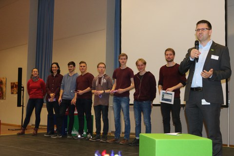 Foto von der Preisverleihung. Studierende stehen in einer Linie vor einer Leinwand. Ein Industrievertreter steht rechts von ihnen und spricht in ein Mikrofon.