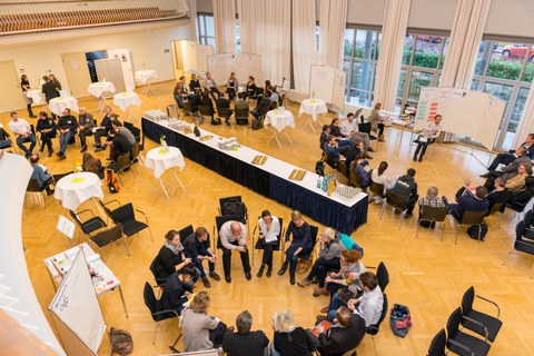 Foto eines großen Saales von oben. In vier großen Stuhlkreisen sitzen Menschen und unterhalten sich. In der Mitte des Saales ist ein Buffet aufgebaut.