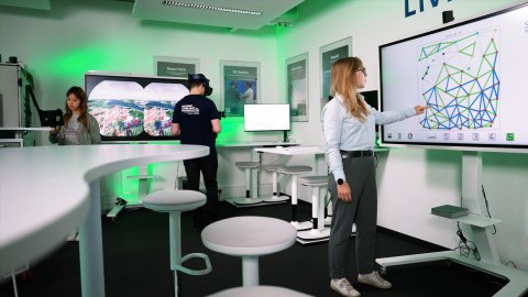 Ein Mann trägt eine Virtual-Reality-Brille, eine Frau arbeitet an einem Tablet, und eine andere Frau zeigt auf eine interaktive Karte auf einem großen Bildschirm. Sie befinden sich alle in einem modernen Büro.