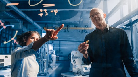Zwei Wissenschaftler, ein Mann und eine Frau, stehen vor einer transparenten Tafel mit Gleichungen. Die Frau zeigt mit dem Finger auf die Tafel, während der Mann sie ansieht. 