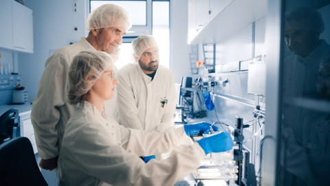 Drei Forschende in Laborkitteln und Kopfbedeckungen arbeiten an wissenschaftlichen Geräten in einem sauberen, modernen Labor.