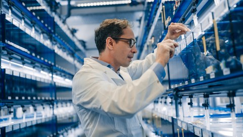 A man wearing a white lab coat and glasses stands in a laboratory inspecting tanks. He reaches for one of the tanks.