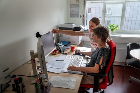 Eine Schülerin des physiotherapeutischen Lehrzentrums zeigt einer Mitarbeiterin  am Schreibtisch den richtigen Abstand zum Bildschirm