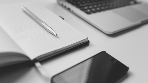 Foto: Arbeitsplatz in schwarzweiß mit einem Handy, Notizbuch samt Stift und einem offenen Laptop.