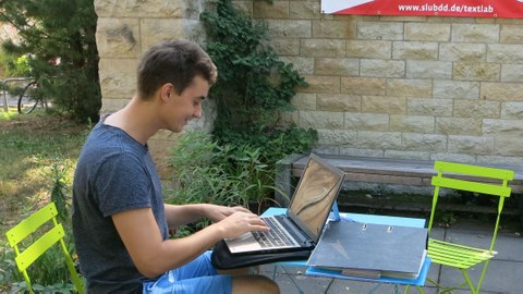 A student sits at an outdoor table in front of his laptop and types. In the background a wall with a banner with: "www.slubdd.de/textlab".