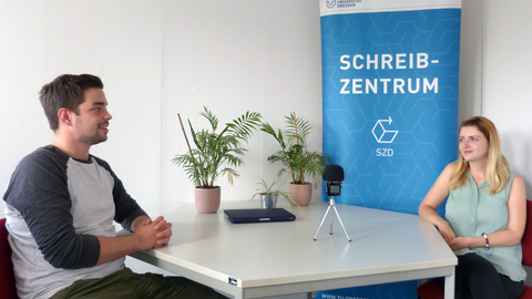 The photo shows two people smiling at each other, sitting at a table in front of an audio recording device. On the table are three green plants, behind them a banner with the logos and lettering of the Writing Centre and the TU Dresden.