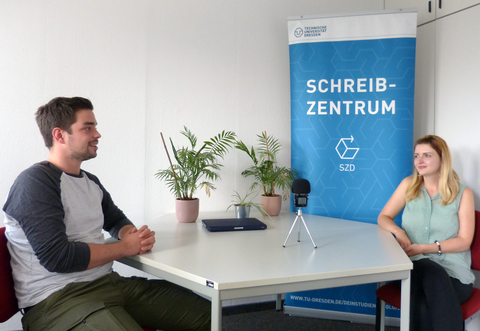 The photo shows two people smiling at each other, sitting at a table in front of an audio recording device. On the table are three green plants, behind them a banner with the logos and lettering of the Writing Centre and the TU Dresden.