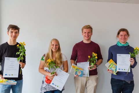 Robin Wratil, Marina Hüttich, Johannes Reimer und Jochen Diepelt schauen lachend in die Kamera. Sie halten in ihren Händen eine gelbe Blume, eine Urkunde und ein Geschenk. Von links nach rechts stehen nebeneinander:  