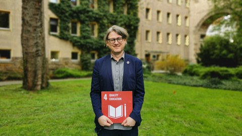 Mann im Hof des Hülße-Bau mit Schild zum SDG 4