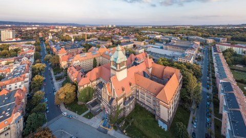 Das Foto zeigt eine Luftaufnahme des Georg-Schumann-Baus der TU Dresden. Das Gebäude wirkt wie eine Art Burg. Besonders auffällig sind der große Wehrturm und die spitzen Dächer. 