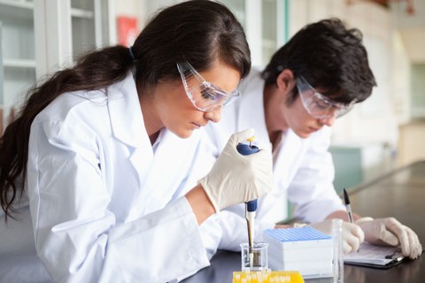 The photo shows two people working in a lab. They are dressed in lab coats and are wearing protective glasses and gloves.