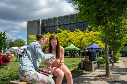 Foto von zwei Frauen, die draußen auf der Bank sitzen und in einen Flyer schauen. Im Hintergrund sieht man verschiedene Stände eines Festes auf dem Campus.