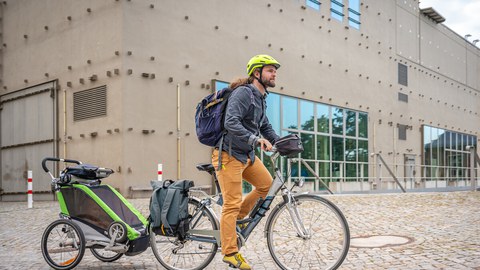 Photo of a young father on a bicycle, riding across the TU campus with a child in the back of a bicycle trailer. 