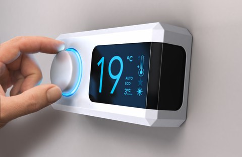 Das Foto zeigt eine Hand, die am Rad eines Thermostates dreht, das an der Wand hängt. Das Thermostat zeigt eine Temperatur von 19 Grad Celsius an.