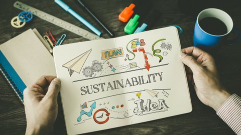 Foto: Ein Arbeitsplatz, über dem zwei Hände ein Notizbuch halten mit einer Skizze zum Thema "Sustainabilty".