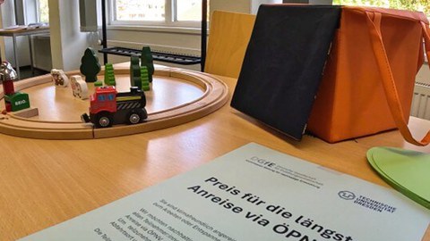 Vorn liegt ein Zettel mit dem Hinweis, dass ein Preis für die längste Anfahrt mit der Bahn vergeben wird. Im Hintergrund ist eine kleine Holzeisenbahn.