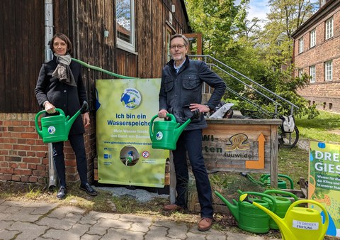 Zwei Personen stehen mit grünen Gießkannen vor einem Wasserbehälter, um daraus Wasser in die Kannen abzufüllen.