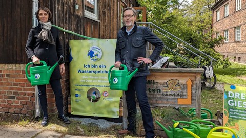 Zwei Personen stehen mit grünen Gießkannen vor einem Wasserbehälter, um daraus Wasser in die Kannen abzufüllen.