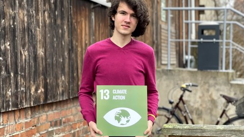 Ein Mann der eine grüne Karte mit weißer Schrift in den Händen hält mit der Aufschrift 13 Climate Action und darunter ein Symbol eines Auges mit der Landkarte drin. Im Hintergrund sieht man den Eingang der Stura Baracke. 