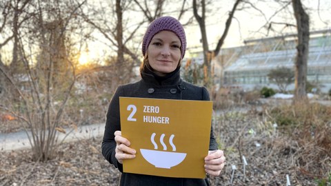Eine Frau der eine gelbe Karte mit der weißen Aufschrift 2 Zero Hunger in den Händen hält. Im Hintergrund sind Sträucher.