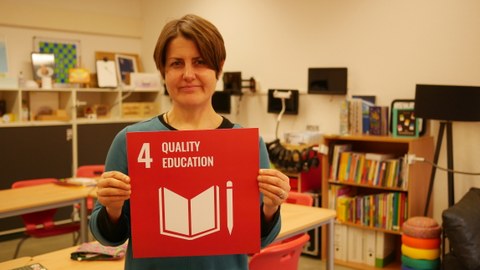 Eine Frau die eine rote Karte mit der weißen Aufschrift 4 Quality Education in den Händen hält. Im Hintergrund ist ein Kursraum.