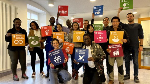 Viele Frauen und Männer die bunte Karten in den Händen halten, welche die Themen der SDG beinhalten und jeweils mit einem Symbol untersetzt sind. 