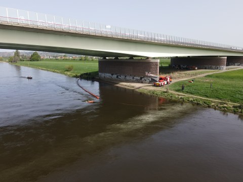 Foto einer Brücke über einen breiten Fluss. Neben deren Pfeiler steht eine Feuerwehr, darum Feuerwehrleute. auf dem Fluß schwebt ein orangefarbener Schlauch mit einem kugelförmigen Ende.