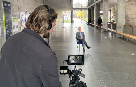 Foto eines Mannes, der im Hörsaalzentrum der TU Dresden eine Videokamera auf einem Stativ bedient. Im Bild ist außerdem ein weiterer Mann abgebildet, der auf einem Stuhl sitzt und gefilmt wird.