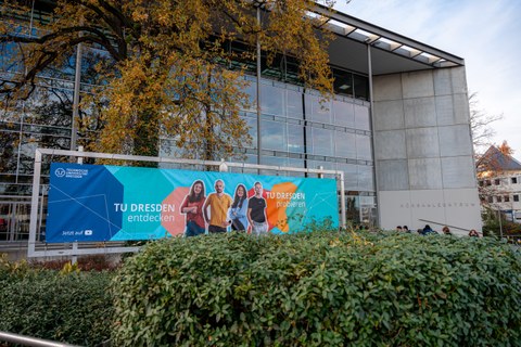Fotoaufnahme des Hörsaalzentrums der TU Dresden. Im Vordergrund ist ein Banner abgebildet, das vier Personen zeigt und die Slogans "TU Dresden entdecken" und "TU Dresden probieren" beinhaltet.