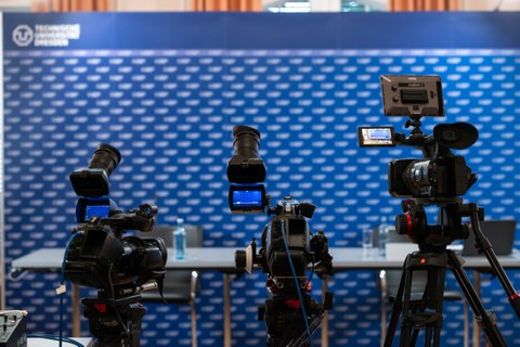 Fotoaufnahme von drei Videokameras, die auf eine blaue Pressewand mit TU Dresden-Logo gerichtet sind. Vor der Pressewand befinden sich zwei Tische und zwei Stühle.