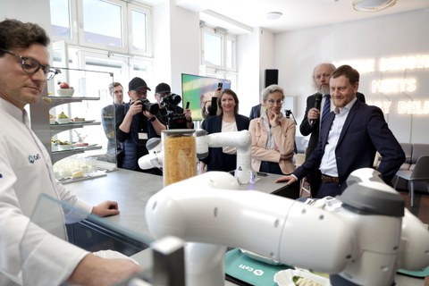 Im Vordergrund ein Mann im weißen Kittel an einer Bar, auf der rechts ein weißer Roboterarm zu sehen ist. Links davon ein hohes Glas, das mit gelben Flocken gefüllt ist, daneben eine Glasvitrine mit Speisen. Vor der Bar stehen v.l. der Sächsische Ministerpräsident, Prof. Fitzek, die Rektorin und andere Personen.