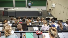 Foto von oben: Ein Dozent schreibt an der Tafel in einem Hörsaal mit aufmerksamen Studenten.