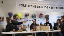 Mehrere Personen sitzen um einen Tisch. Darauf befinden sich Essen, Getränke, und Modellfiguren. Im Hintergrund hängen Masken und ein Banner auf dem "Politische Bildung" steht.