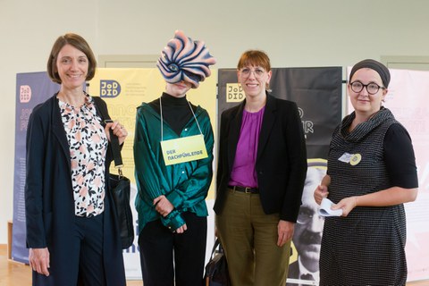 Vier Frauen lächeln in die Kamera, eine davon trägt eine Maske.