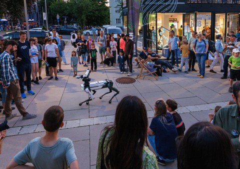 Draußen vor einem Gebäude steht eine große Gruppe Menschen im Kreis um einen vierbeinigen Roboter, der sich in der Mitte des Platzes bewegt. Kinder und Erwachsene schauen interessiert zu, einige filmen oder fotografieren die Szene. Die Atmosphäre ist neugierig und entspannt.