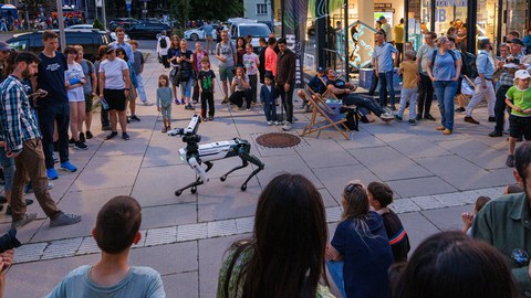 Draußen vor einem Gebäude steht eine große Gruppe Menschen im Kreis um einen vierbeinigen Roboter, der sich in der Mitte des Platzes bewegt. Kinder und Erwachsene schauen interessiert zu, einige filmen oder fotografieren die Szene. Die Atmosphäre ist neugierig und entspannt.